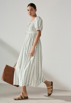 Frau in einem weißen Kleid mit vertikalen grünen Blumenstreifen, die eine braune geflochtene Tasche hält, beige Sandalen trägt und goldene Creolenohrringe trägt.