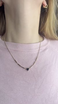 Colar dourado com elos alongados e pendente em forma de coração preto. Modelo a usar um suéter cor-de-rosa claro; foco nos detalhes do colar.
