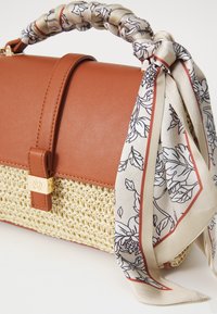Brown leather handbag with a woven straw base and a patterned cream scarf tied around the handle, featuring gold hardware accents.