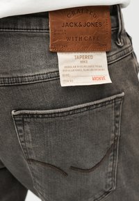 Καφέ δερμάτινο ταμπελάκι και υφασμάτινη ετικέτα στην τσέπη γκρι τζιν με λεπτομέρειες της μάρκας Jacks Jones και περιγραφή του στενού Mike fit.