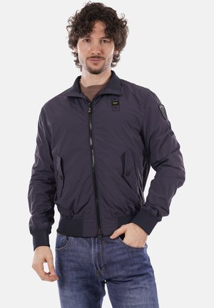 Uomo con capelli ricci che indossa una giacca blu navy con cerniera e jeans blu, in piedi con una mano in tasca su sfondo bianco.