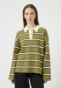 Kvinne med våt-look hår som bærer perleøreringer og en olivengrønn stripete genser med hvit krage og beige knapper.