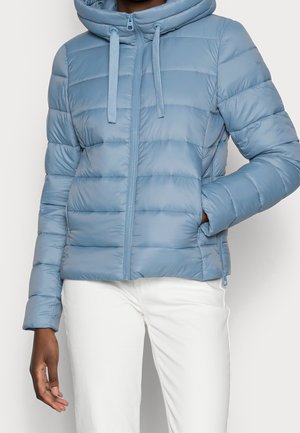 Lichtblauwe gewatteerde jas met capuchon en ritssluiting aan de voorkant, gedragen met een witte broek, één hand in de zak en de andere ontspannen langs het lichaam.