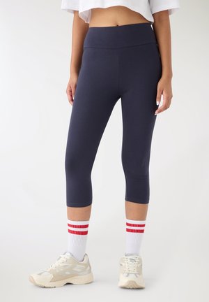 Granatowe, wysokowaistowe leginsy capri wykonane z elastycznego materiału, zestawione z białymi skarpetkami z czerwonymi paskami oraz beżowymi butami sportowymi.