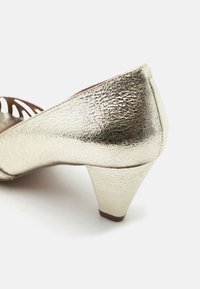 Παπούτσι με χαμηλό τακούνι χρυσού χρώματος με υφή και σχέδιο με αποκοπές κοντά στα δάχτυλα, εμφανίζεται από το πλάι σε λευκό φόντο.