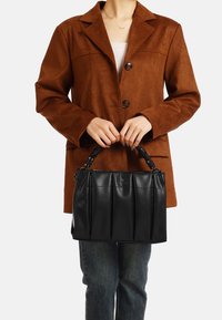 Borsa a mano in pelle nera con design plissettato e manico intrecciato, tenuta davanti a una persona che indossa una giacca in suede marrone e jeans.