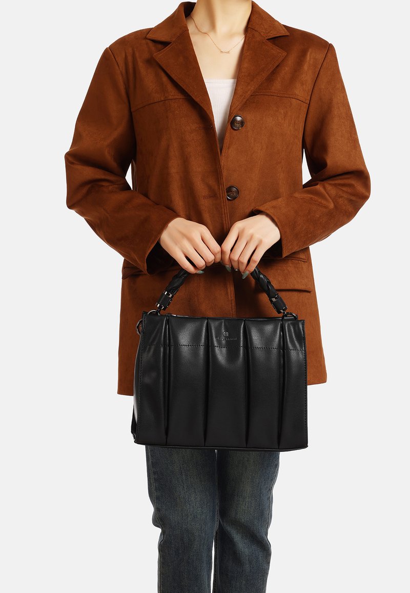 Borsa a mano in pelle nera con design plissettato e manico intrecciato, tenuta davanti a una persona che indossa una giacca in suede marrone e jeans.
