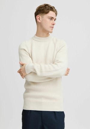 Ribbed-knit cream sweater with a high neck, featuring long sleeves and a fitted silhouette, paired with dark navy trousers.