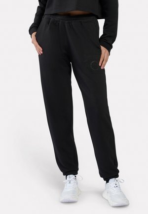 Persoon draagt zwarte joggerbroek met elastische boorden en witte sneakers, staat met handen in de zakken.