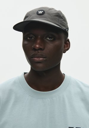 Mladić tamnije puti u svijetloplavoj majici i sivoj kapici s bijelim logotipom "W", okrenut prema naprijed na jednobojnoj pozadini.