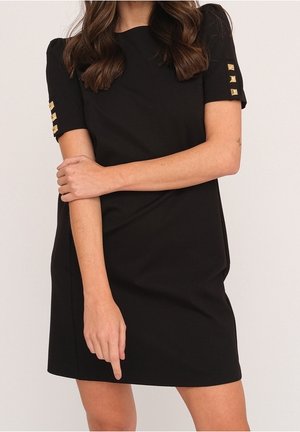 Kvinde iført en sort kjole med korte ærmer og guld firkantede nitter på ærmerne, stående med armene krydset og pegende nedad med fingeren.