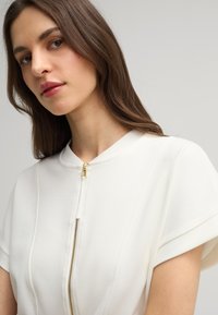 Witte top met een korte rits bij de kraag, voorzien van gestructureerde schouders en een zachte textuur. Gouden ritsaccent. Stijlvol, minimalistisch ontwerp.