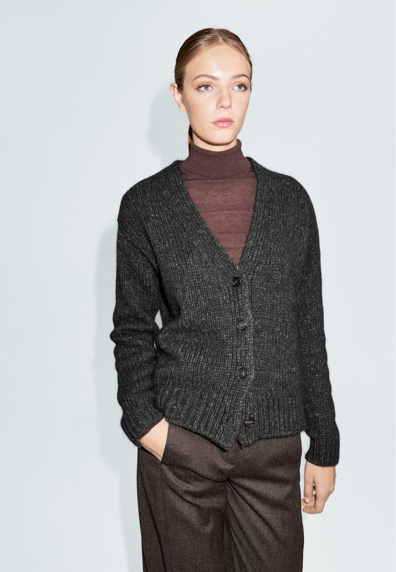 Donkergrijze gebreide cardigan met V-hals, knoopsluiting en ribbelstructuur. Gelaagd over een bruine coltrui, gecombineerd met een paarse broek.