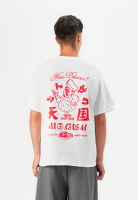 T-shirt en coton blanc avec un imprimé graphique rouge au dos, représentant un chef, du texte et un graphique de pizza. Manches courtes, coupe décontractée.