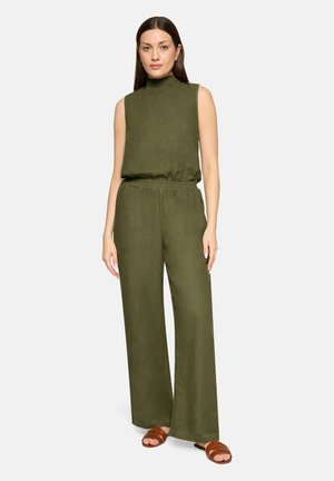 Kobieta stojąca, ubrana w oliwkowozielony, bezrękawnik z wysokim kołnierzem i pasujące szerokie spodnie oraz brązowe sandały na jasnym tle.