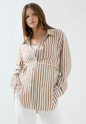 Nainen, jolla on suorat ruskeat hiukset, pukeutunut beigeen ja valkoiseen pystysuoraiseen raidalliseen paitaan valkoisen topin ja valkoisten housujen päällä, kädet taskuissa.