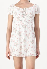 Nainen, jolla on yllään valkoinen, lyhyt, kukallinen mekko, jossa on pinkkejä kukkia, puffihihat ja nauhakiinnitys pääntiellä.