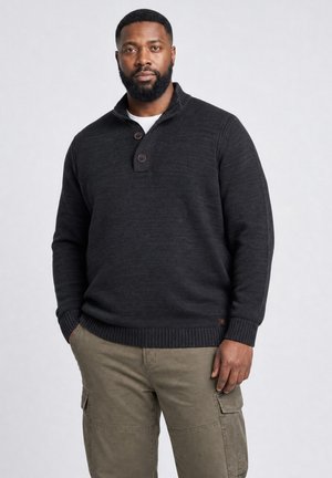 Mann mit kurzen schwarzen Haaren und Bart, der einen dunklen, geknöpften Pullover und khakifarbene Cargohosen trägt, steht mit einer Hand in der Tasche vor einem einfarbigen Hintergrund.