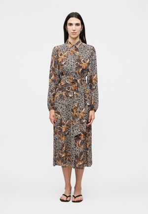 Žena má na sebe midi šaty s dlhým rukávom a golierom s hnedým, čiernym a bielym kvetinovým a abstraktným vzorom, doplnené čiernymi sandálmi.