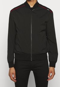 Giacca bomber nera con chiusura a zip, polsini e orlo a coste, e strisce rosse sui fianchi. Realizzata in tessuto leggero e liscio.