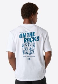 Mand vender ryggen iført en hvid T-shirt med blå tekst "Du kan finde mig PÅ KLIPPENE" og tegneserieisterninger, der danser på ryggen.