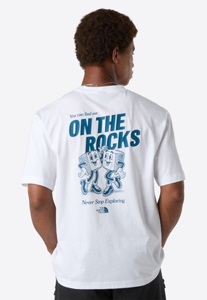 Mand vender ryggen iført en hvid T-shirt med blå tekst "Du kan finde mig PÅ KLIPPENE" og tegneserieisterninger, der danser på ryggen.