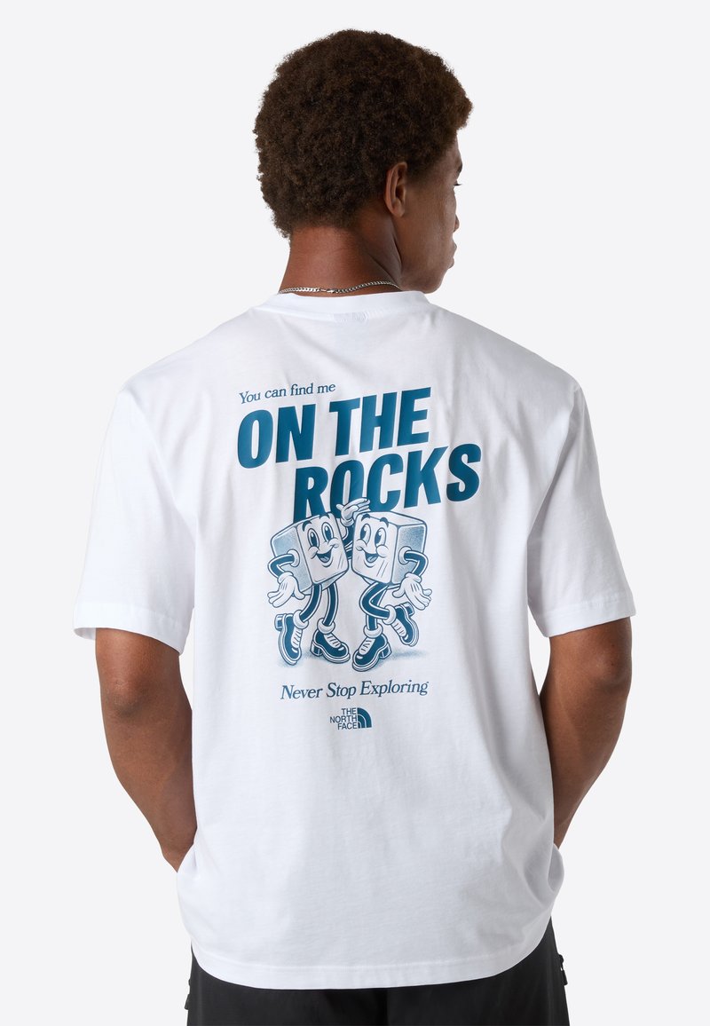 Mand vender ryggen iført en hvid T-shirt med blå tekst "Du kan finde mig PÅ KLIPPENE" og tegneserieisterninger, der danser på ryggen.
