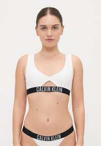 Bralette blanc à côtes avec un design découpé, bande logo noire 'CALVIN KLEIN', bas de bikini assortis avec une taille noire.