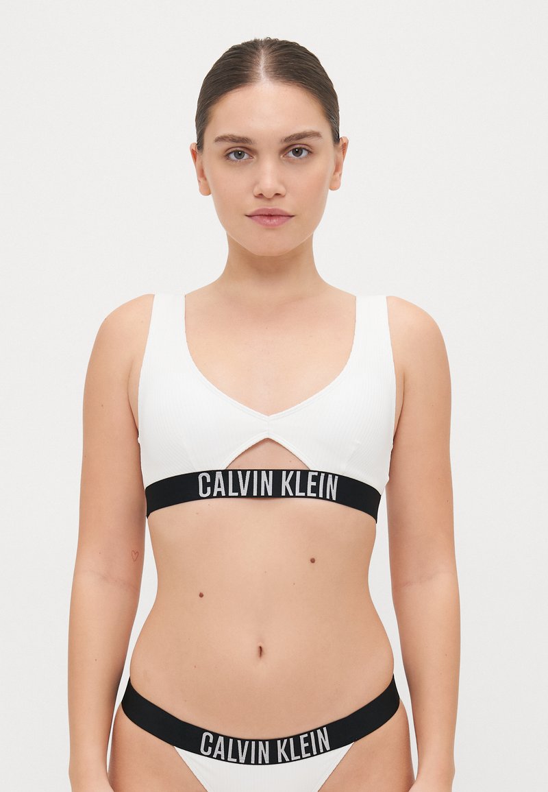 Bralette blanc à côtes avec un design découpé, bande logo noire 'CALVIN KLEIN', bas de bikini assortis avec une taille noire.