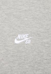 Siva tkanina puloverja z nežno teksturo. V zgornjem desnem kotu je bela vezenina z logotipom Nike SB.
