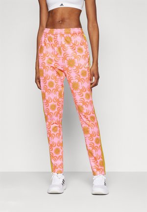 Kvinde iført pink og orange mønstrede træningsbukser, hvid sports-bh og hvide Adidas-sneakers, stående mod en ensfarvet baggrund.
