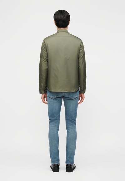 Homme portant une chemise à boutons vert olive, un jean bleu clair et des chaussures noires, debout de dos sur un fond uni.