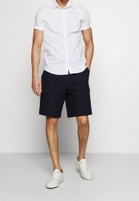 Vit kortärmad skjorta med knappar och en subtil logotyp, i kombination med marinblå texturerade shorts och vita sneakers, vilket skapar en avslappnad outfit.