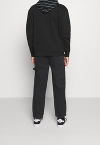 Svart sweatshirt med huva som har blå ränder, i kombination med svarta cargobyxor och svarta sneakers med vita detaljer, sedd bakifrån.