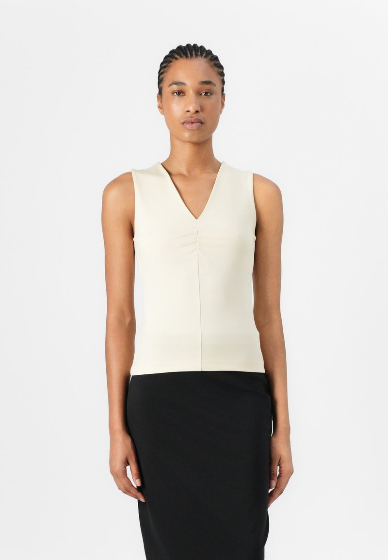 Beige ärmelloses Top mit V-Ausschnitt und gerafftem Detail in der Mitte, kombiniert mit einem schwarzen Bleistiftrock, glattes Material und figurbetonter Schnitt.