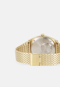 Montre-bracelet dorée avec un bracelet en mesh extensible. Boîtier rectangulaire avec un dos poli et une couronne pour le réglage. Marquée "TIMEX."