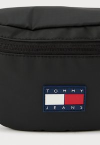 Zwarte pouch gemaakt van glad, gestructureerd materiaal. Heeft een ritssluiting en een blauwe label met witte en rode strepen, met de tekst "TOMMY JEANS."