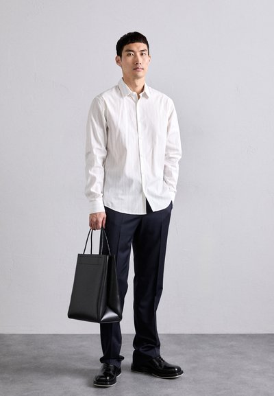 Camisa blanca a rayas finas, pantalones oscuros, bolsa de cuero negra con asas, zapatos negros, de pie contra un fondo gris claro.