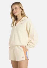 Πουλόβερ κρεμ χρώματος με γιακά μισού φερμουάρ, σετ με σορτς στο ίδιο χρώμα που έχουν πορτοκαλί και μπλε πλευρικές λεπτομέρειες. Μαλακό, υφασμάτινο ύφασμα με υφή.