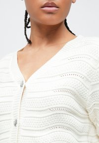 Γυναίκα με πλεξούδες που φοράει λευκό πλεκτό ζακετάκι με διακοσμητικά ασημένια κουμπιά, παρουσιάζοντας υφή με κυματιστά σχέδια στο ύφασμα.
