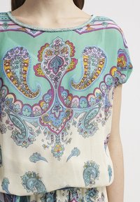 Kevyt paita, jossa on turkoosi pohja ja monimutkaisia paisley- ja kukkakuvioita violetilla, keltaisella ja sinisellä. Helmillä koristeltu kaula-aukko.