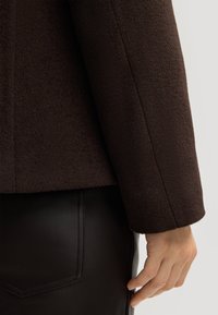 Manica di giacca a trama marrone abbinata a un indumento in pelle nera, che mette in evidenza linee pulite e una silhouette aderente.