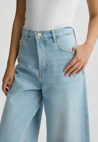 Jeans de culoare albastru deschis, cu picioare largi și talie înaltă, având un închideri cu buton aurit, buzunare frontale și o ușoară decolorare pentru textură.