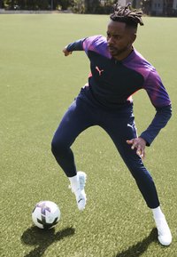 Puma Sportoutfit in Marineblau mit rosa Akzenten, bestehend aus einer Zip-Jacke und eng anliegenden Hosen. Weiße Sportschuhe und ein Schwarz-Weiß Fußball.