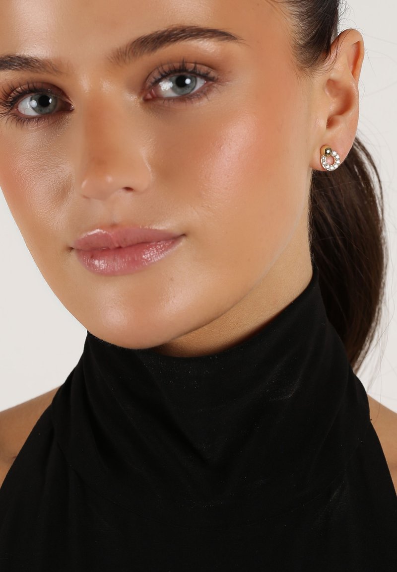 Silver earrings with a circular design, featuring clear rhinestones and a gold accent. Worn on a model with a black high-neck top.
