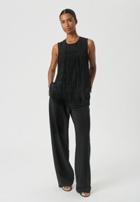 Črn brezrokavnik s teksturiranim detajlom franj na sprednji strani, v kombinaciji z ohlapnimi črnimi širokimi hlačami in črnimi ravnimi sandaali.