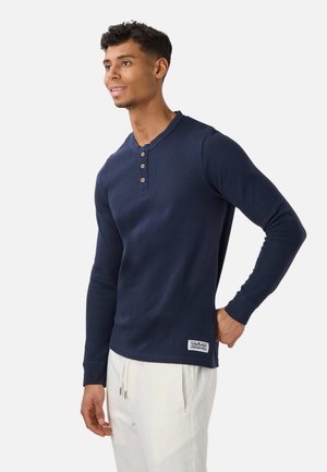 Młody mężczyzna w granatowej bluzce henley z długim rękawem i guzikami oraz białych spodniach ze sznurkiem, stoi z jedną ręką na biodrze.