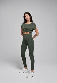 Olijfgroen cropped top met knoop aan de voorkant, gecombineerd met bijpassende high-waisted leggings. Model draagt witte sportschoenen. Fluweelachtige textuur.