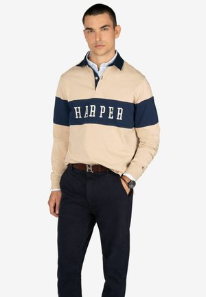 Polo a maniche lunghe beige con una striscia blu navy, con la parola "HARPER" in bianco. Abbinato a pantaloni blu navy scuri e una cintura marrone.