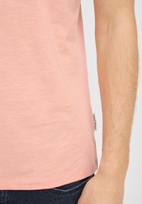 Camiseta de algodón rosa claro con dobladillo redondeado, que presenta una pequeña etiqueta en el lateral. La tela tiene una textura suave y un sutil brillo.